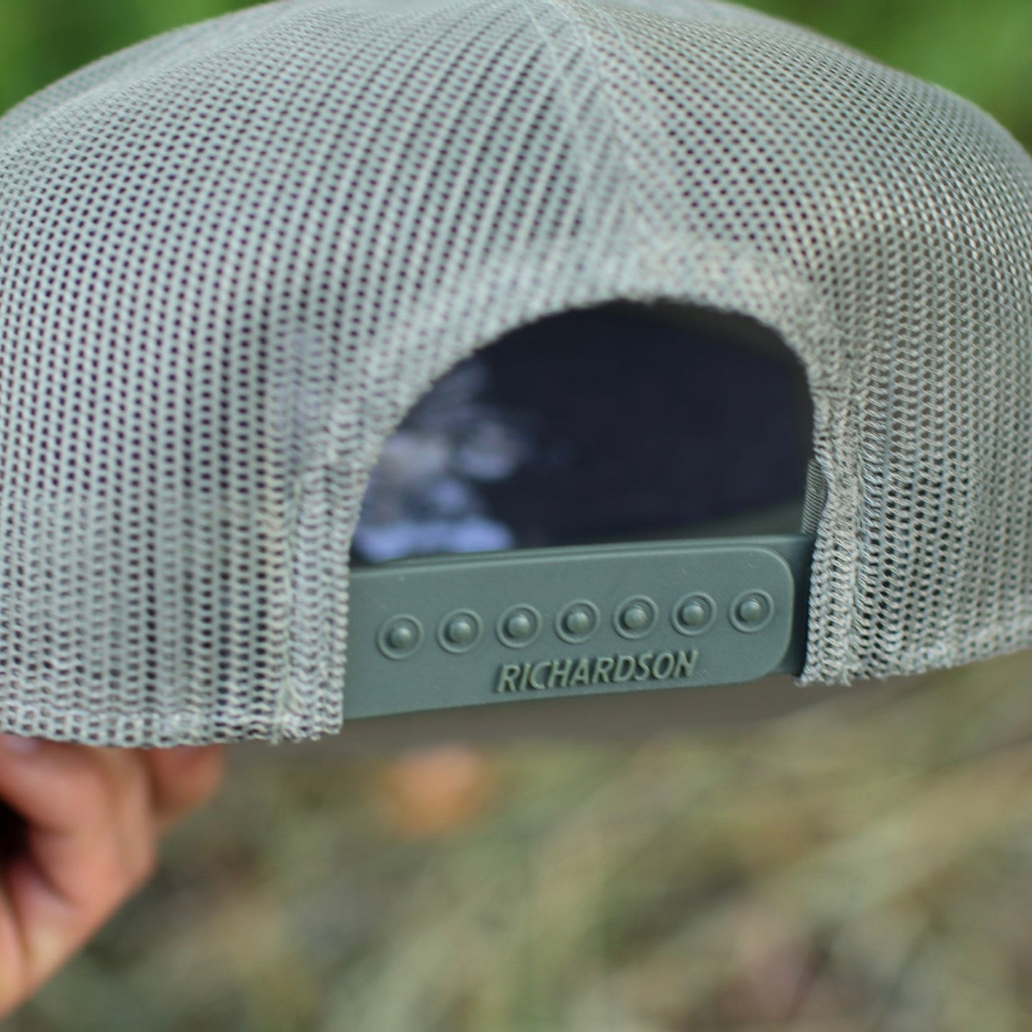 Close-up of a gray mesh cap with 'Richardson' branding, held against a blurred natural background.