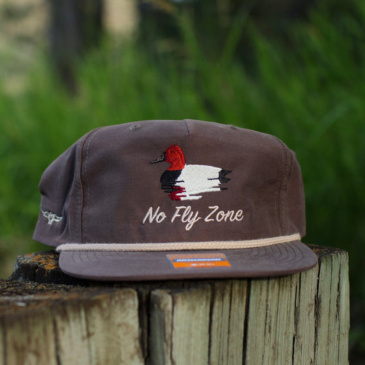 Brown cap with 'No Fly Zone' text and duck design of a canvasback on a wooden post with a blurred green background