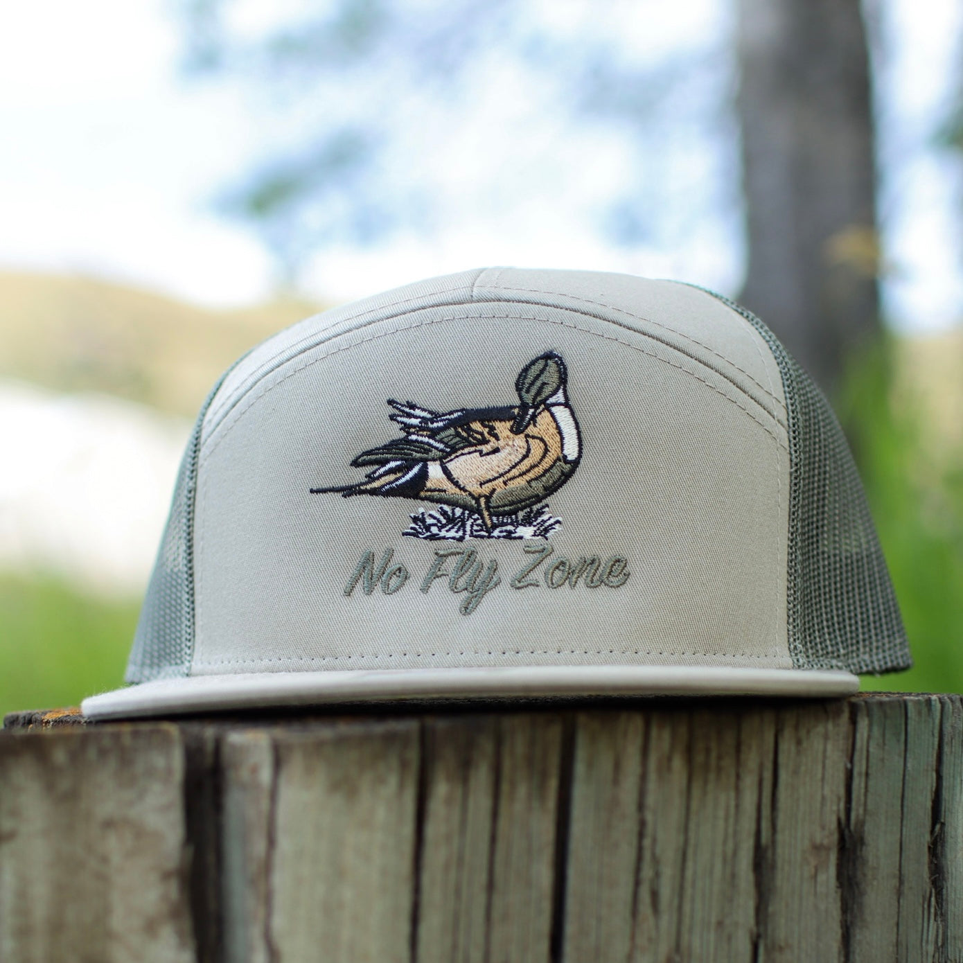 Trucker hat with duck design and 'No Fly Zone' text on a wooden post with a blurred natural background