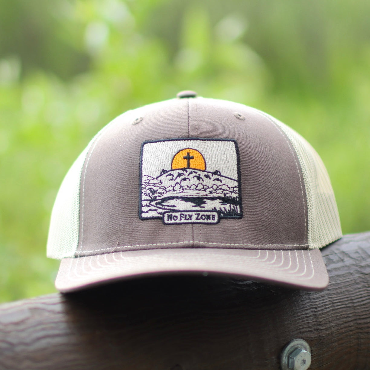 Gray cap with a patch featuring a sunset and mountains with a cross on a blurred green background.