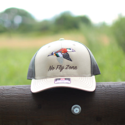 Cap with 'No Fly Zone' text and wigeon design on a wooden surface with a blurred natural background